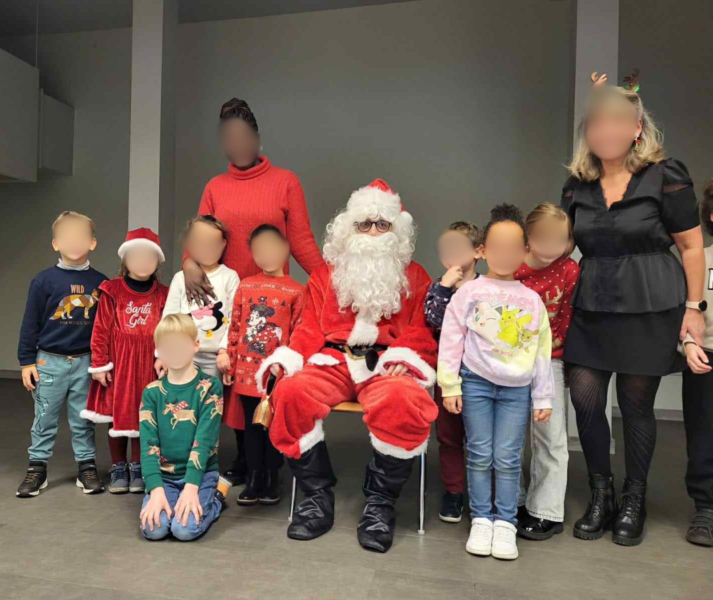 Père Noël assis entouré d’enfants et de deux adultes lors d’un moment festif de fin d’année en intérieur.