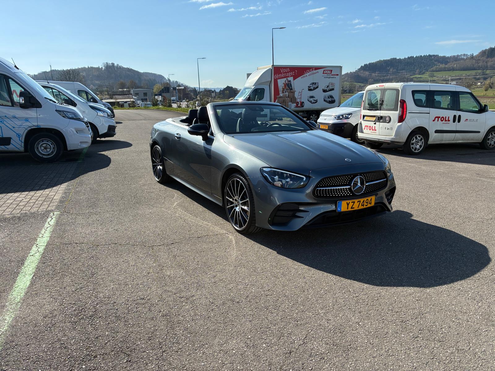 Voiture cabriolet grise sur parking ensoleillé.