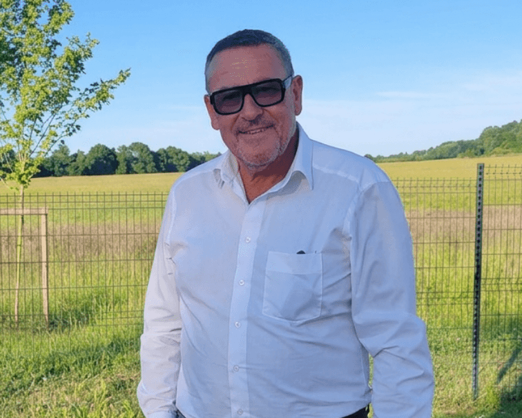 Mann mat Sonnebrëll virun engem Feld a jonk Bam
