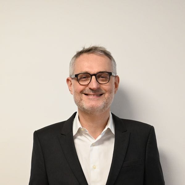 Homme souriant en costume noir devant mur blanc