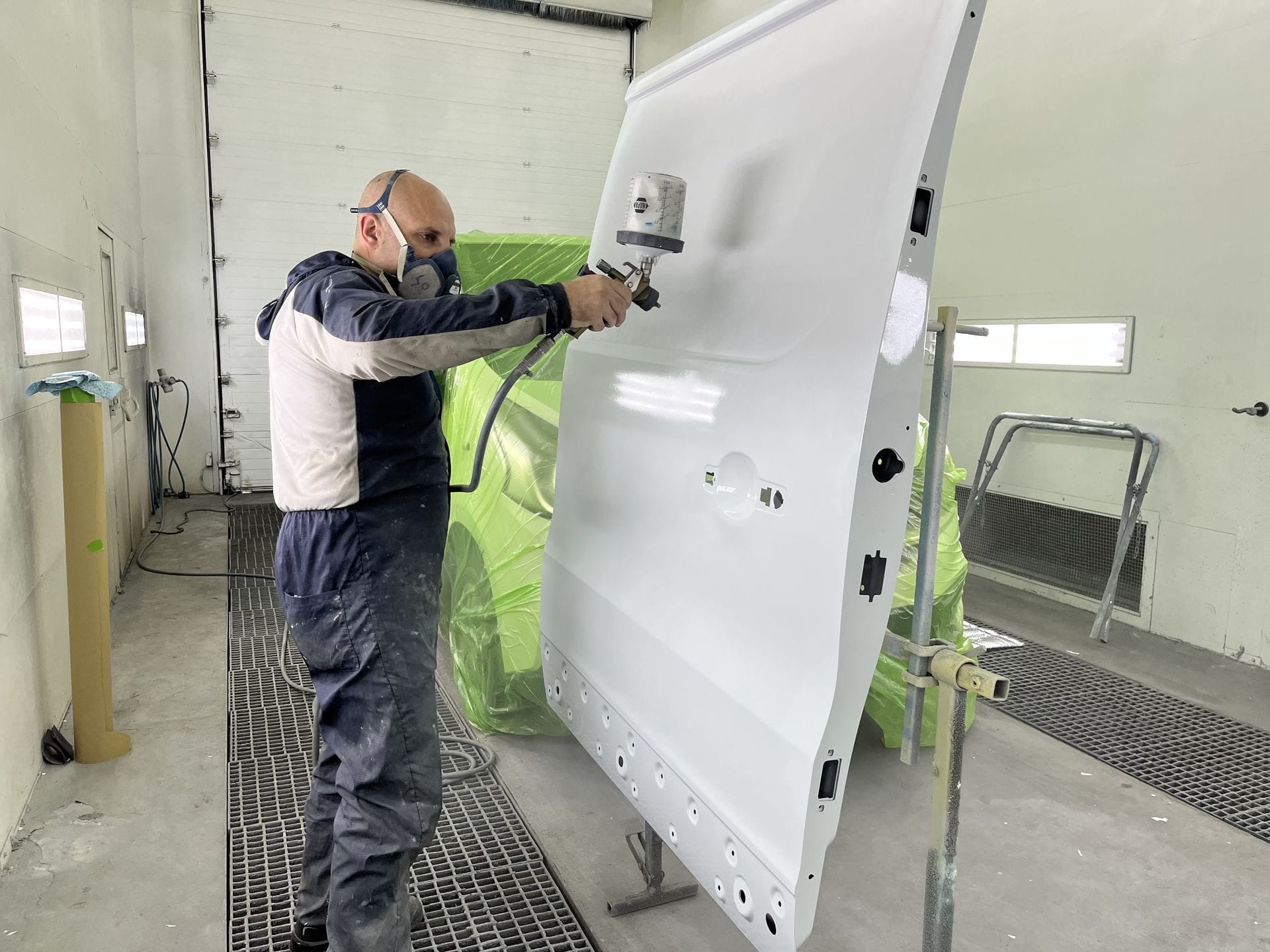 Un technicien en blouse et masque applique une couche de peinture blanche sur une portière de véhicule utilitaire.
